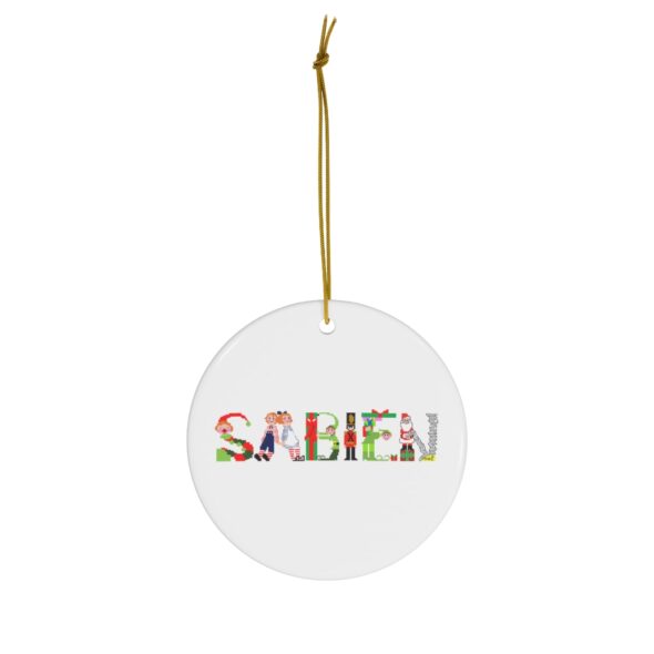 White ceramic ornament with text ‘Sabien’ in colourful Christmas themed lettering, with gold hanging loop